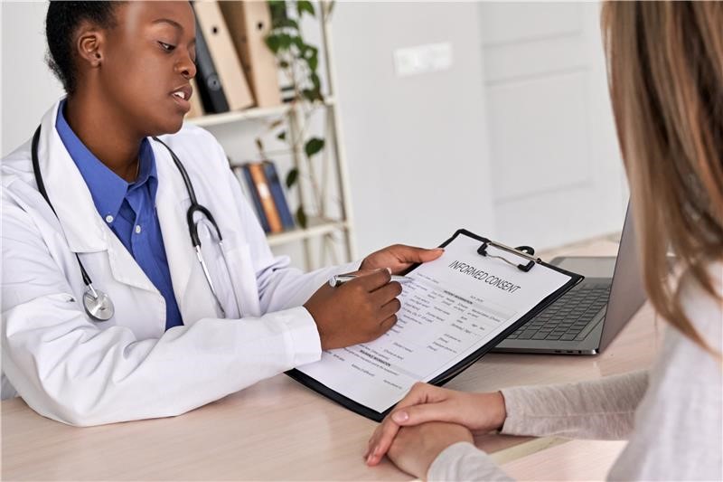 Clinician and patient sitting across from each other at a desk. The clinician is holding a clipboard with a paper that reads Informed Content at the top. The patient has her hands folded together as she listens to the clinician explain the document and signature requirements.