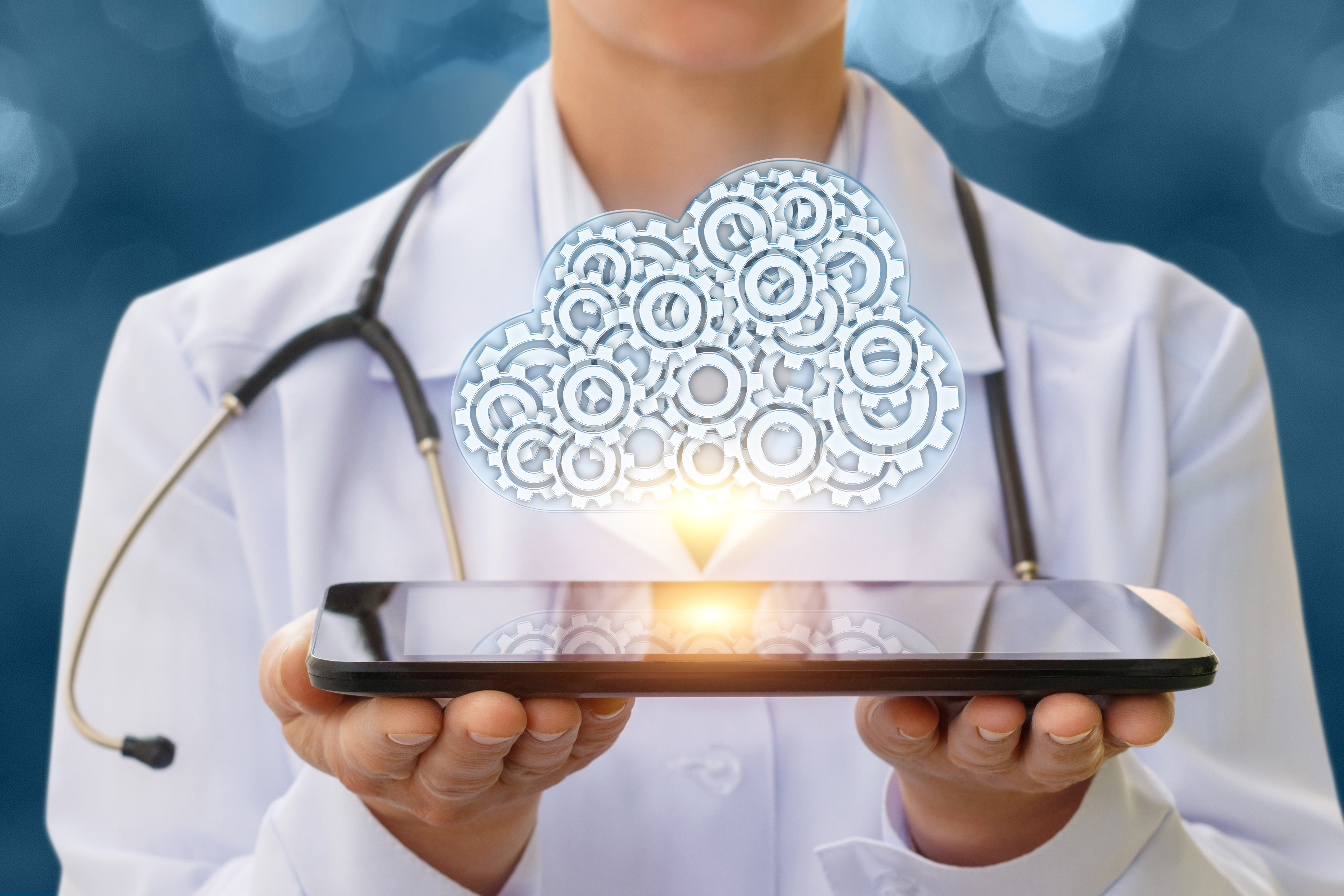 A person in a white lab coat, wearing a stethoscope, holds an electronic tablet. Above the tablet is the image of a cloud, indicating a place where data are sent for storage.