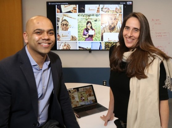 Drs. Nikhil Wagle (on the left) and Corrie Painter (on the right) lean against a table with a laptop in between them. Behind them in a large computer monitor presenting information about “Count Me In” that includes picutes of participants in the program.