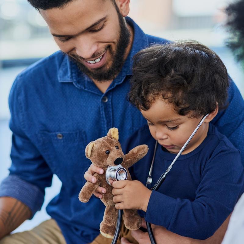 Male child uses stethoscope to listen for teddy bear's pulse. Child sits on adult male's lap.