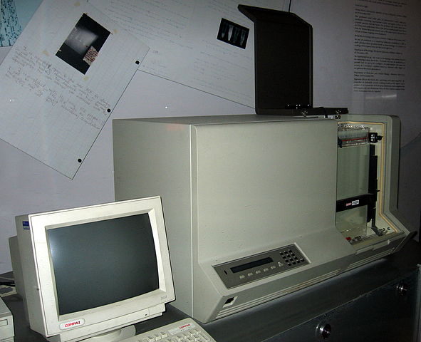The first automated DNA sequencer sitting on a desk, Compaq box monitor off to the left with CPU on the right. Setting is a dated office space with papers tacked to the wall, muted dingy wall color.
