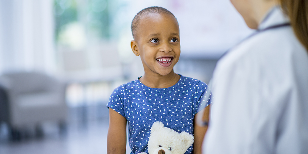 Photo portrays a small child in a blue dress sitting and holding a stuffed teddy bear and talking with a doctor. Although she has cancer, as shown by her cancer-related hair loss, she’s happy and smiling at her doctor. 