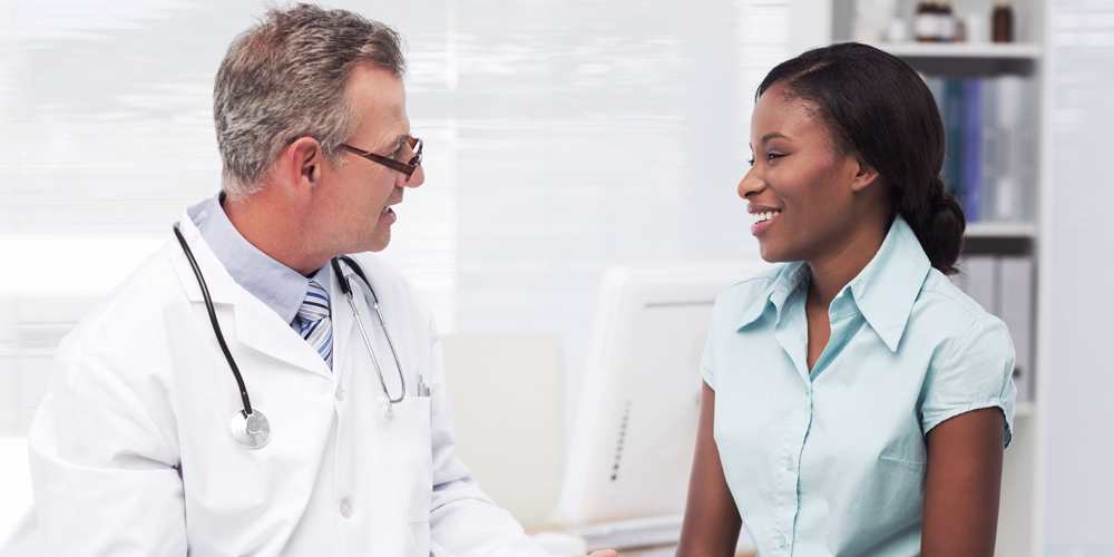 Photo of a white male doctor sitting next to a young African American woman. The doctor is speaking to the woman and the woman is looking at him and smiling. 