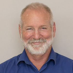 Headshot of Michael McNitt, man with gray beard smiling at the camera. Blue shirt. 
