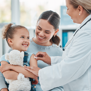 Image of a doctor looking at a young girl holding a stuffed animal while her mother holds her.