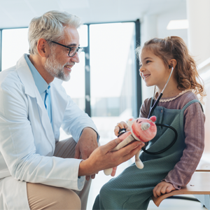 doctor looking at child in medical setting. the child has stethescope in ears and the doctor holds a stuffed animal out to the child. Both are smiling. 