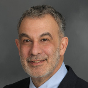Professional headshot of Joel Saltz, M.D., Ph.D. Dr. Saltz is dressed up, smiling, and looking directly at the camera. The background is a dark grey color.