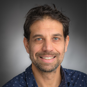 Headshot of a man with a blue collared shirt and grey background.