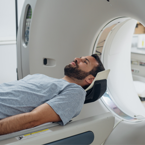 Man dressed in a hospital gown and laying on his back. He is being transferred into a CT scanning machine.