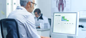 Photo of a male scientist carefully examining findings on his desktop computer screen. His hands are on the keyboard and he is entering information. Another scientist sits in the background and a poster showing human anatomy hangs on the wall.