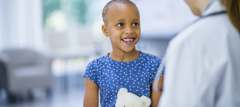 Photo portrays a small child in a blue dress sitting and holding a stuffed teddy bear and talking with a doctor. Although she has cancer, as shown by her cancer-related hair loss, she’s happy and smiling at her doctor. 