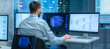 Photo shows a male sitting and facing a row of computer screens. He has his hand resting on the desk and he’s reading the screen that’s immediately in front of him.  