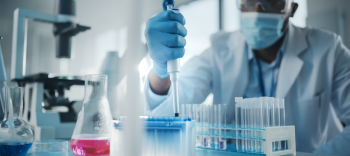 Photo shows a scientist in a laboratory setting. The scientist is wearing a surgical mask and goggles and he is using a syringe to carefully measure liquid into a row of test tubes. 